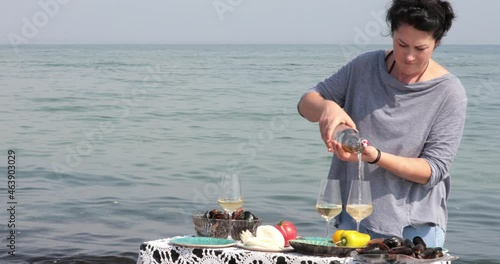 Woman pour white wine into wineglasses, then two middle aged women cheers and drinking wine together at sea water background. Table setting with seafood mussels. Summer beach picnic at seaside photo