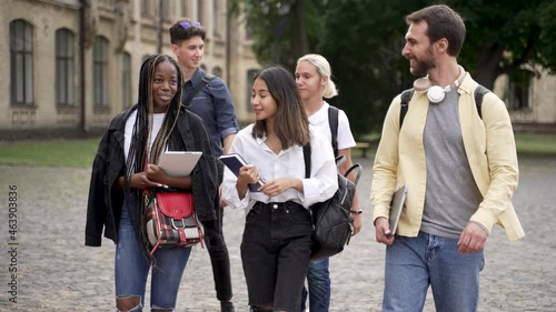 Cheerful multinational high school students chatting while walking through campus park before classes. Positive multi-ethnic friends communicating while going along university park before lessons