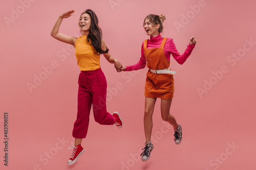 Two interracial young women in their 20s are running on pink background holding hands. Full-length image of dancing girls in bright colors. Leisure concept youth, active lifestyle