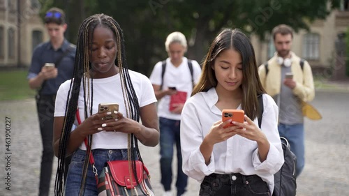 Group of multi-ethnic high school students addicted to smartphones walking through campus park, ignoring everything around them. Obsessed young people phubbing each other using gadgets