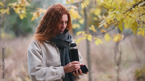 beautiful red-haired woman drinks tea from thermos in the autumnal forest photo