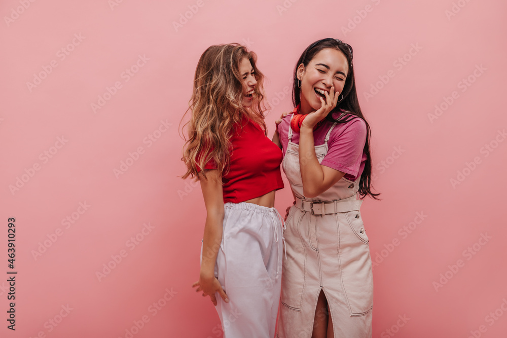 Cheerful two interracial young women are having fun and dancing on pink background. Ukrainian blonde and asian brunette laugh with closed eyes huddled together with happy faces and smiles. Cheerful two interracial young women are having fun and dancing on pink background. Ukrainian blonde and asian brunette laugh with closed eyes huddled together with happy faces and smiles.