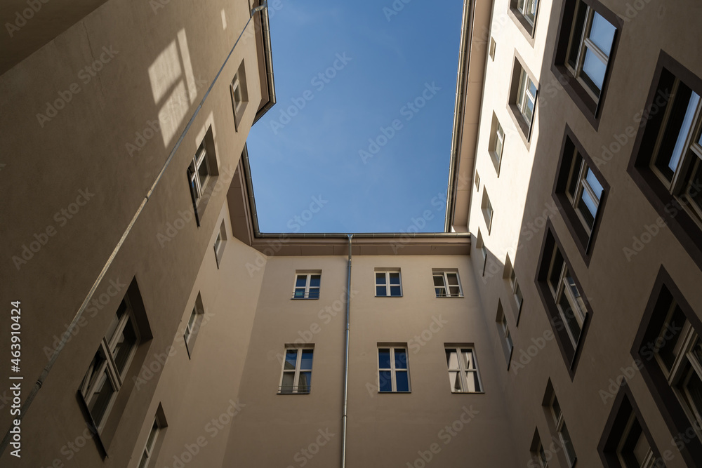 Looking up between tall buildings to the blue sky. Facades of houses in front of the viewer. Sunlight is hitting the wall and windows are reflecting the light. A dark place in the backyard.