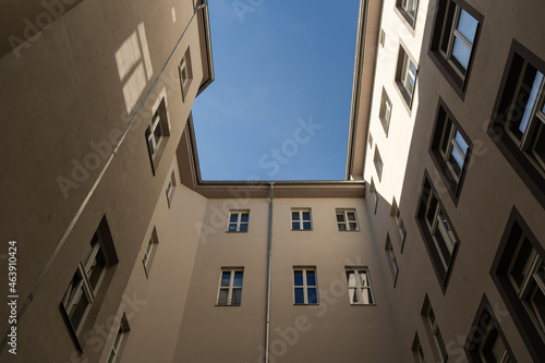 Looking up between tall buildings to the blue sky. Facades of houses in front of the viewer. Sunlight is hitting the wall and windows are reflecting the light. A dark place in the backyard. © 1take1shot