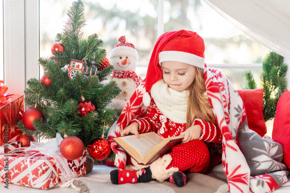 a cute girl child is reading a book sitting on the windowsill at the window of the house at the Christmas tree and waiting for the new year or Christmas in a red Santa Claus hat and smiling a cute girl child is reading a book sitting on the windowsill at the window of the house at the Christmas tree and waiting for the new year or Christmas in a red Santa Claus hat and smiling