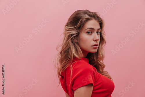 Close-up of elegant young woman staring intently into camera lens against pink background. Girl with loose wavy hair in red T-shirt. Harmony and tranquility concept