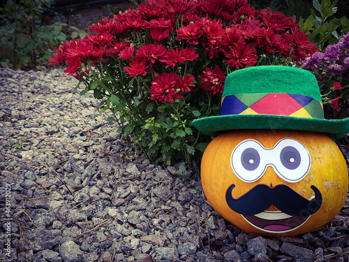 a funny pumpkin with a hat on its head and a mustache stands in the garden. chrysanthemums are blooming in the background. Halloween background