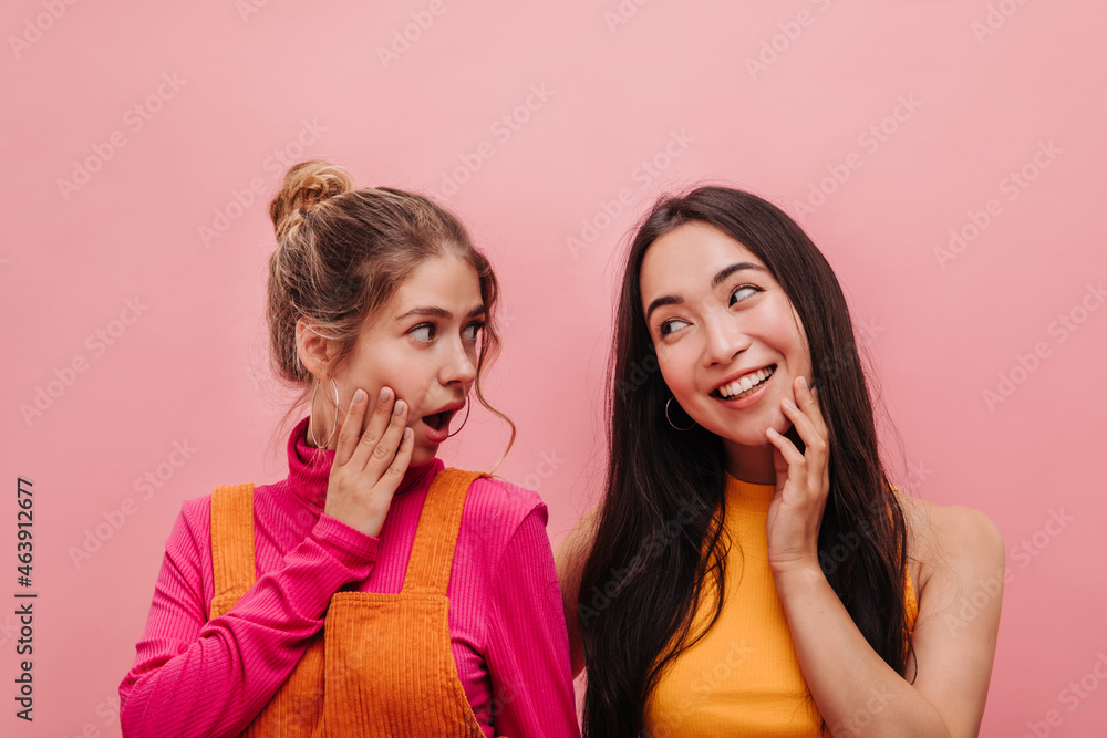 Caucasian young girl is surprised to hear story of asian friend looking at each other. Portrait two best friends, blonde and brunette in autumn casual clothes. Caucasian young girl is surprised to hear story of asian friend looking at each other. Portrait two best friends, blonde and brunette in autumn casual clothes.
