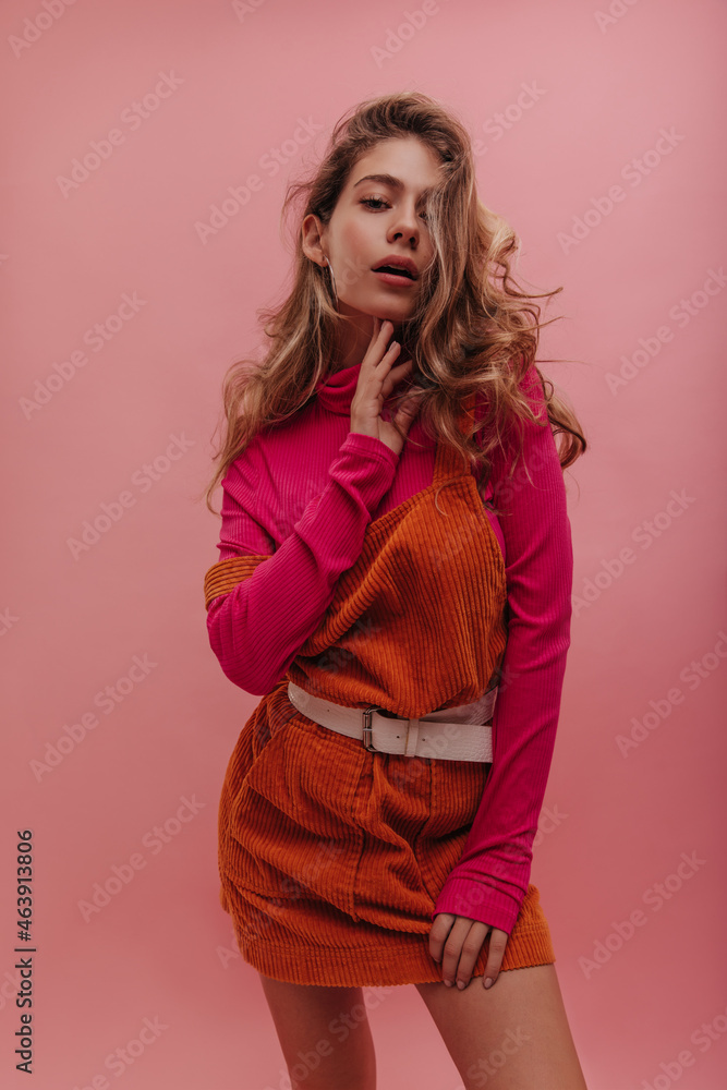 Fototapeta premium Young ukrainian lady posing alone over isolated pink background, thinking about date. Bright outfit of apricot denim dress and sweater, blonde loose curly hair with natural makeup.