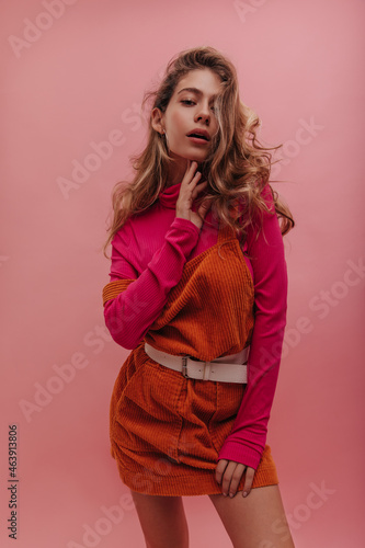 Young ukrainian lady posing alone over isolated pink background, thinking about date. Bright outfit of apricot denim dress and sweater, blonde loose curly hair with natural makeup.