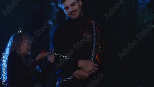 Medium shot of vampire caught in darkness standing at tree in forest as courageous girl tying metal rope locking entity. Halloween party in dark woodland outdoors