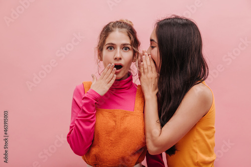 Fotografie Asian brunette woman whispers secret to her friend from Europe, surprising her on pink background with place for text