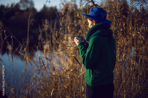 woman in a green knitted sweater and a blue hat stands by the bushes at sunset and drinks tea from an iron thermos mug