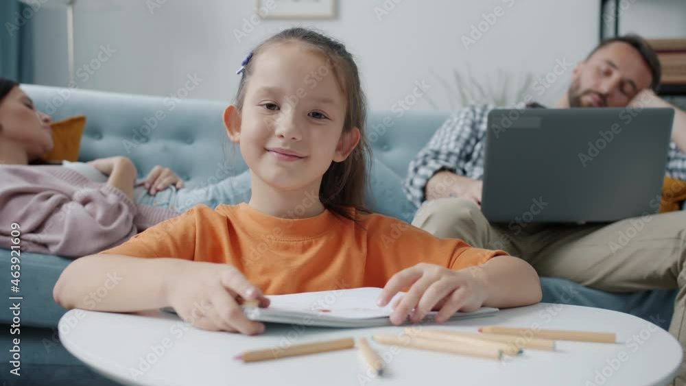 Portrait of cheerful kid smiling holding pencil drawing sitting at table while mom and dad are sleeping in background. Family and lifestyle concept. Portrait of cheerful kid smiling holding pencil drawing sitting at table while mom and dad are sleeping in background. Family and lifestyle concept.