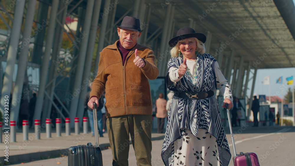 Senior happy pensioner tourists grandmother grandfather with luggage near international airport hall raises thumbs up agrees something or gives positive reply recommends advertisement likes good, ok Senior happy pensioner tourists grandmother grandfather with luggage near international airport hall raises thumbs up agrees something or gives positive reply recommends advertisement likes good, ok