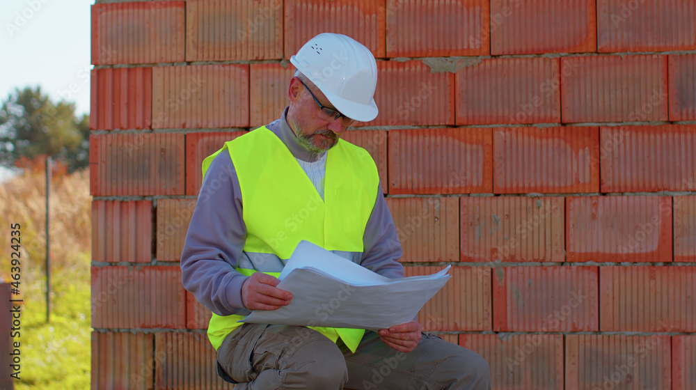 Civil engineer architect specialist analyzing blueprints to control working at construction site. House on background. Developer builder foreman in helmet inspecting building. Real estate home project Civil engineer architect specialist analyzing blueprints to control working at construction site. House on background. Developer builder foreman in helmet inspecting building. Real estate home project