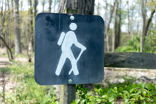 Hand  painted sign with white  paint shows a person with backpack  and walking stick hiking along a path,  mounted  to fence  post at edge of forest trail to mark trailhead entrace