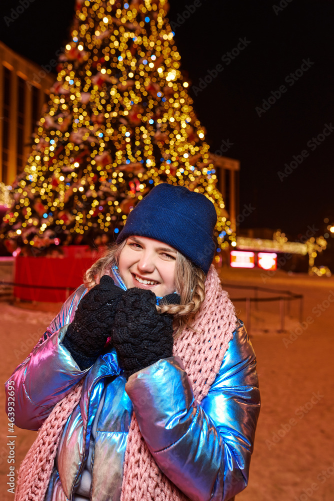 Happy smiling young Caucasian woman in scarf, hat, jacket, mittens by the christmas tree outdoor. New year, fun, winter concept 