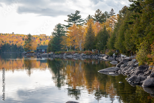 A rocky lakeshore leads to a quaint cabin. The trees have reached peak foliage in this scene. 