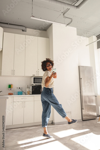 Full-length photo of happy multiethnic female wearing wireless headphones and dancing on the kitchen while holding smartphone © Yaroslav Astakhov