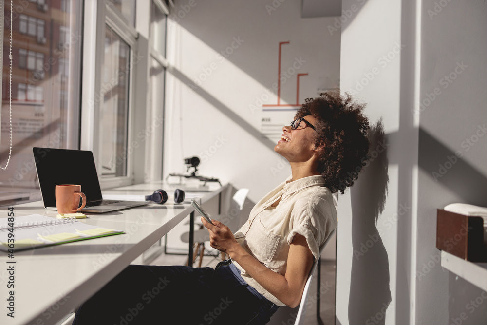 Side view of happy Afro American lady using mobile phone while working in the office near the window Side view of happy Afro American lady using mobile phone while working in the office near the window