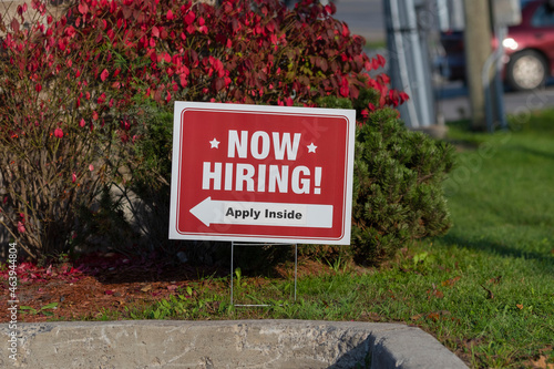 Outdoor lawn sign now hiring apply inside with a direction arrow, selective focus, blurred background. Employment and labor shortage, resigning, great resignation concept. photo