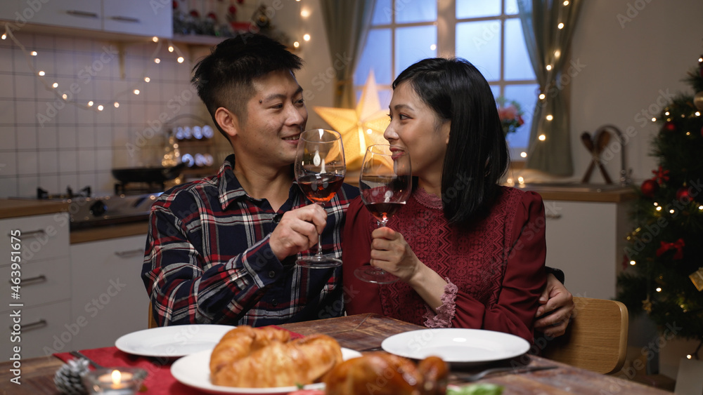 happy asian couple sitting close at dinner table and making toast to celebrate valentine’s day in a romantic home interior with light decors