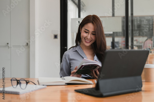 Smiling asian woman reading a book while studying or webinar online via tablet, video conference concept © Natee Meepian