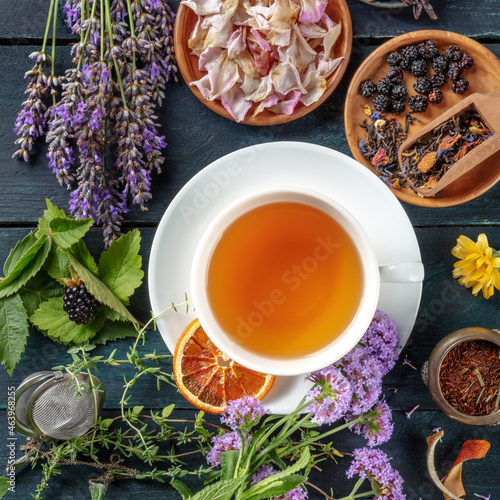 A cup of tea with dry fruit, flowers, and herbs on a dark rustic wooden background, square overhead shot