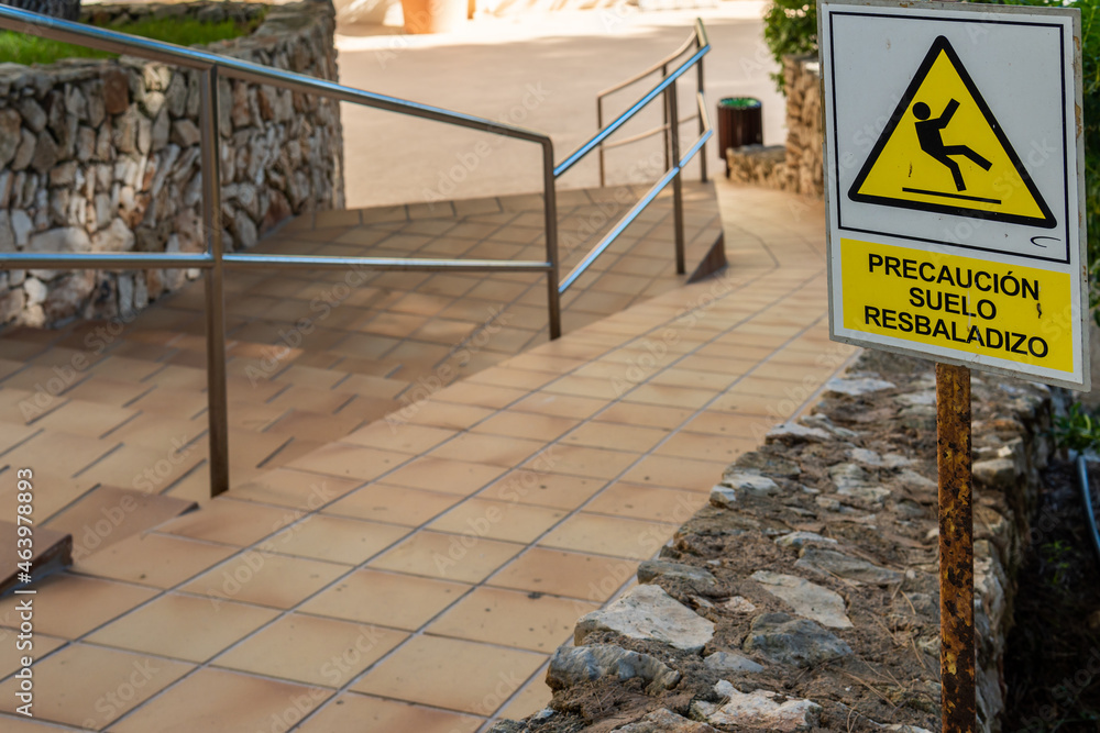 Close-up of the slippery ground caution sign, written in Spanish language. In the background, an access ramp to a hotel complex Close-up of the slippery ground caution sign, written in Spanish language. In the background, an access ramp to a hotel complex