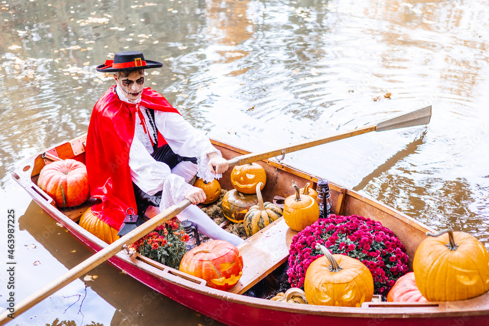 young man dressed as gondolier in gondola boat decorated with pumpkins on pond in autumn park celebrates Halloween and having fun, concept of Halloween carnival or costume party - obrazy, fototapety, plakaty 