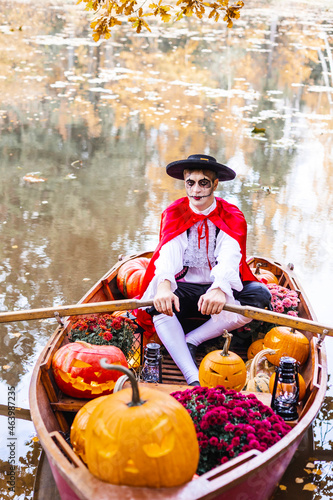 young man dressed as gondolier in gondola boat decorated with pumpkins on pond in autumn park celebrates Halloween and having fun, concept of Halloween carnival or costume party