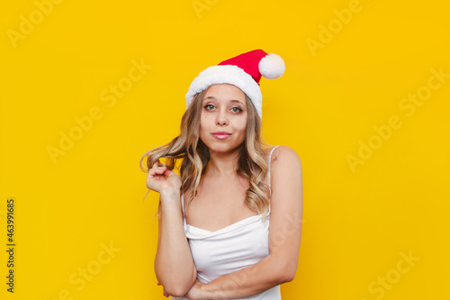 A young caucasian confident sexy slender blonde woman in a white top and a red Santa hat twisting a lock of hair on her finger isolated on a color yellow background. Christmas and New Year concept © Марина Демешко