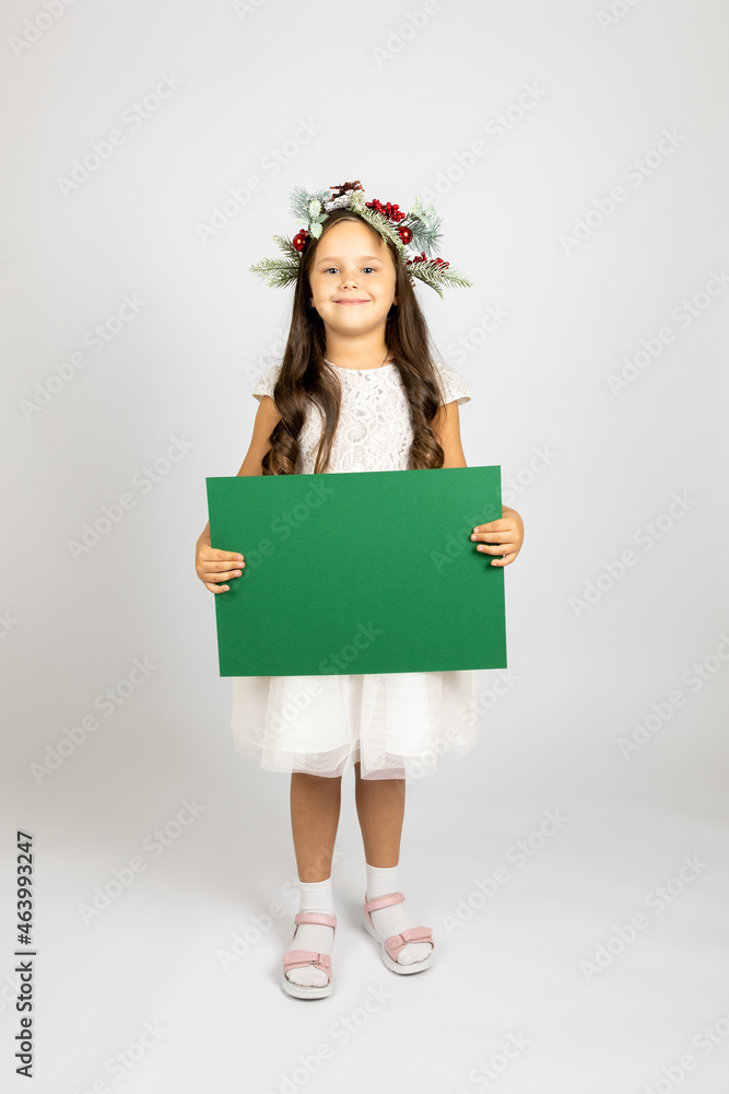full-length portrait of charming preschool-age girl in Christmas wreath and white festive dress holding green empty poster, isolated on white background. full-length portrait of charming preschool-age girl in Christmas wreath and white festive dress holding green empty poster, isolated on white background.