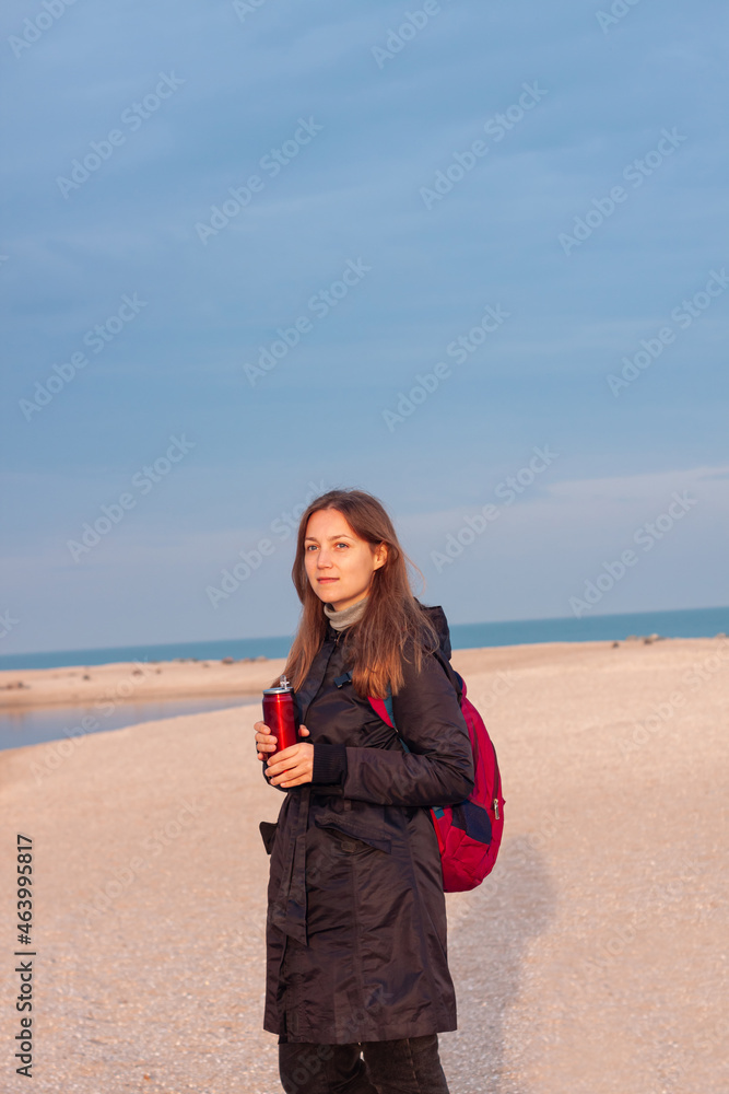 Happy young woman in black raincoat with backpack on empty beach autumn sea. Smiling millennial girl with long hair walking hiking drinking tea in thermo can bottle alone.Lifestyle real people outdoor Happy young woman in black raincoat with backpack on empty beach autumn sea. Smiling millennial girl with long hair walking hiking drinking tea in thermo can bottle alone.Lifestyle real people outdoor