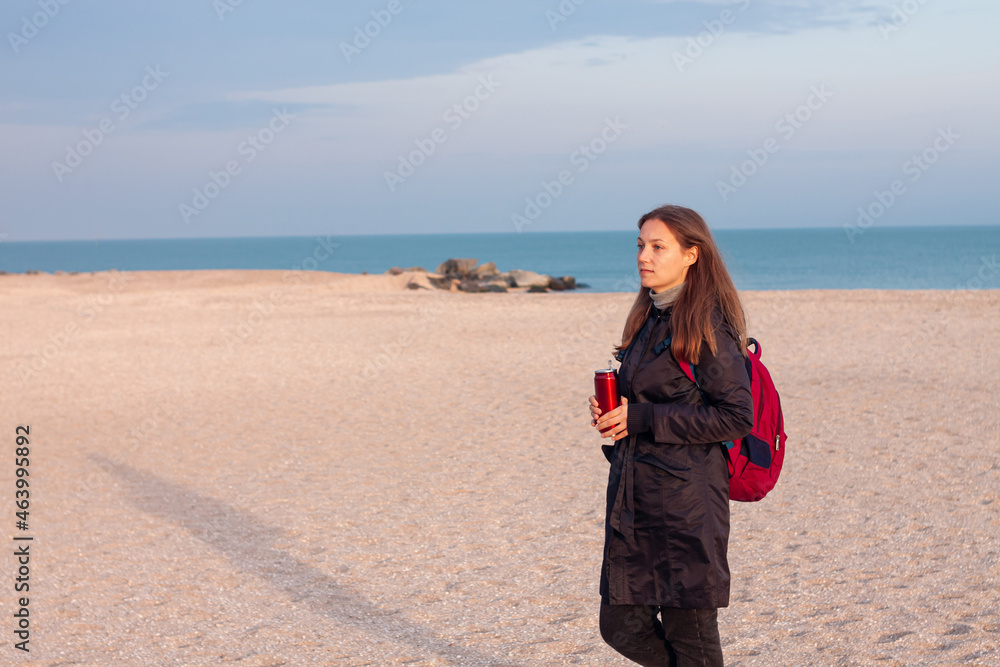 Happy young woman in black raincoat with backpack on empty beach autumn sea. Smiling millennial girl with long hair walking hiking drinking tea in thermo can bottle alone.Lifestyle real people outdoor