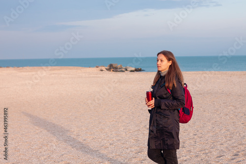 Happy young woman in black raincoat with backpack on empty beach autumn sea. Smiling millennial girl with long hair walking hiking drinking tea in thermo can bottle alone.Lifestyle real people outdoor