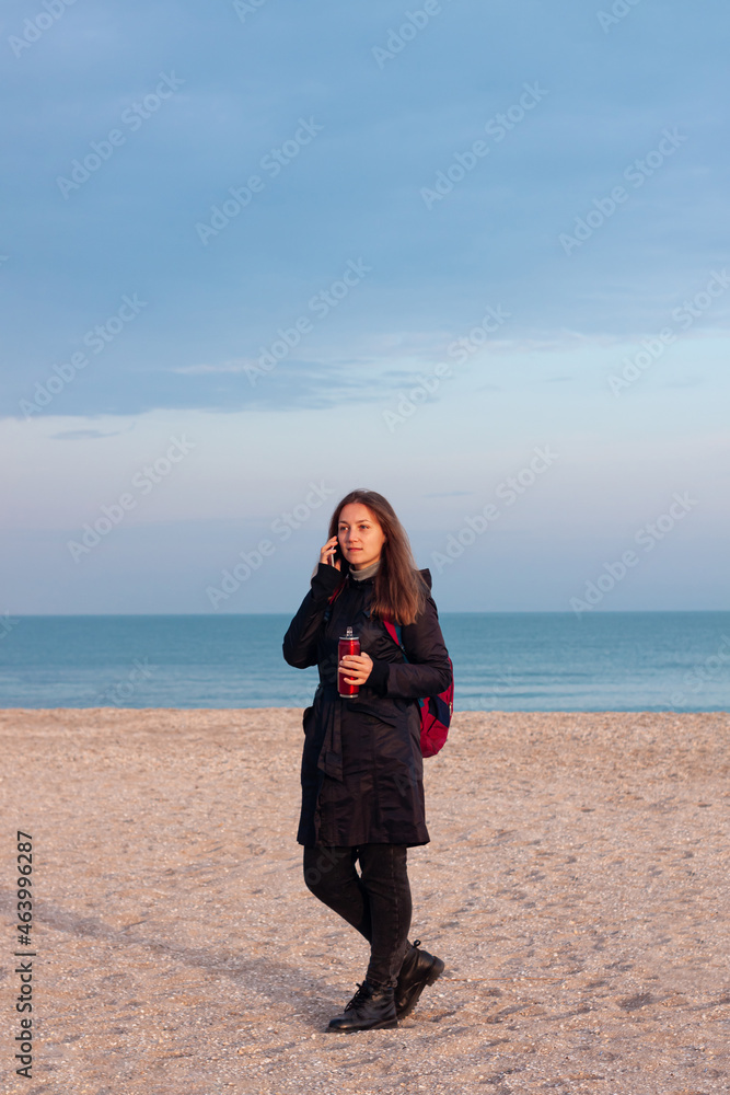 Happy young woman in black raincoat with backpack on empty beach autumn sea. Smiling millennial girl with long hair walking hiking drinking tea in thermo can bottle alone.Lifestyle real people outdoor