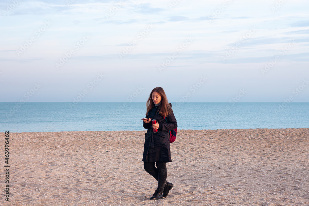 Happy young woman in black raincoat with backpack on empty beach autumn sea. Smiling millennial girl with long hair walking hiking drinking tea in thermo can bottle alone.Lifestyle real people outdoor Happy young woman in black raincoat with backpack on empty beach autumn sea. Smiling millennial girl with long hair walking hiking drinking tea in thermo can bottle alone.Lifestyle real people outdoor