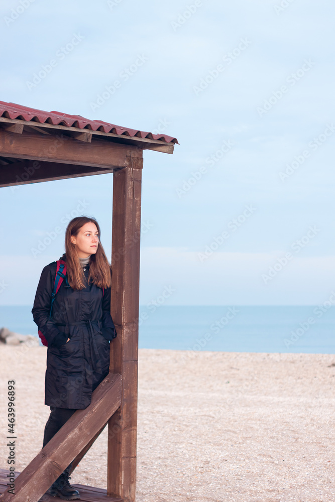 Happy pretty young woman in black raincoat with backpack on empty beach autumn sea. Smiling millennial girl with long hair walking hiking resting alone. Lifestyle real people female portrait outdoor