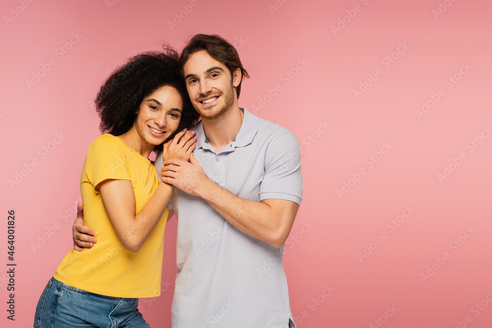 young and happy multiethnic couple embracing while smiling at camera isolated on pink. young and happy multiethnic couple embracing while smiling at camera isolated on pink.