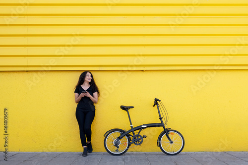 Young black woman with long hair and black outfit texting  somebody next to her Bicycle In Front Of A Yellow Wall