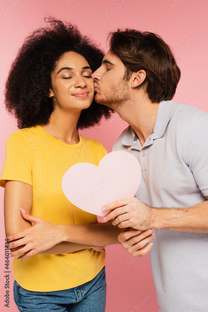 man with paper cut heart kissing happy hispanic girlfriend isolated on pink.