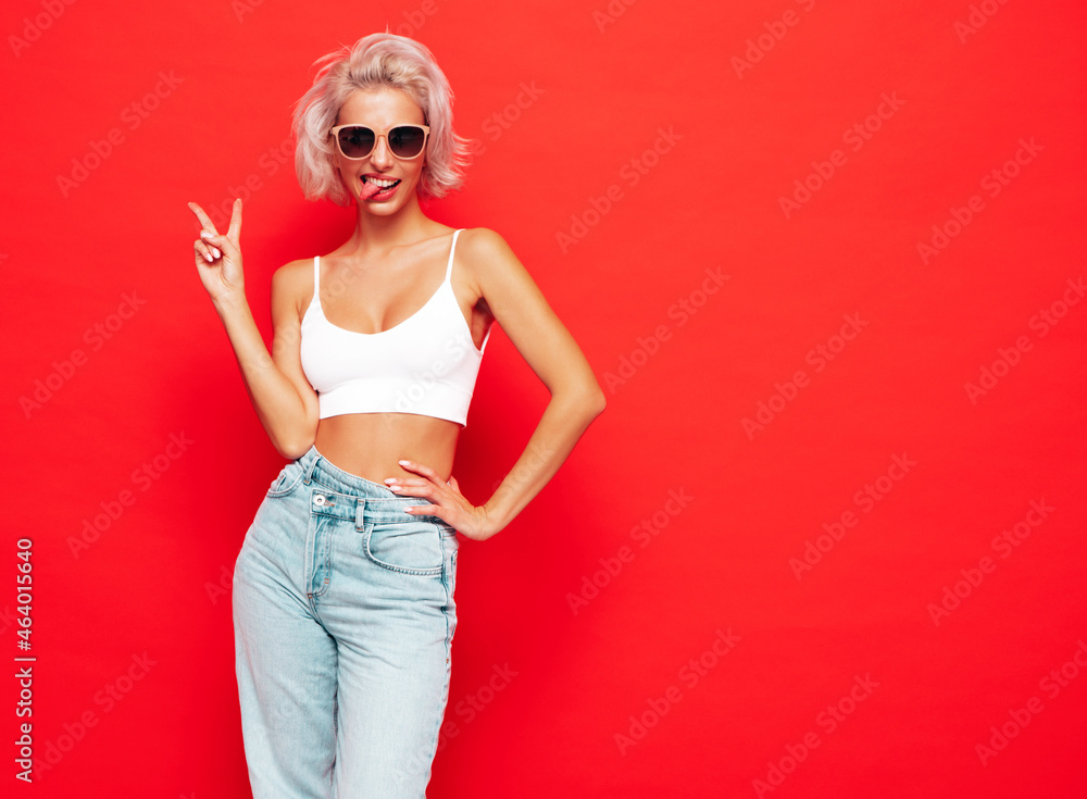 Young beautiful smiling female in trendy summer clothes. Sexy carefree woman posing near red wall in studio. Positive blond model having fun and going crazy. Cheerful and happy. In sunglasses Young beautiful smiling female in trendy summer clothes. Sexy carefree woman posing near red wall in studio. Positive blond model having fun and going crazy. Cheerful and happy. In sunglasses