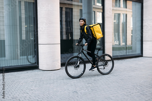 Smiling courier girl riding a bicycle wearing delivery thermal backpack looking for a customer address