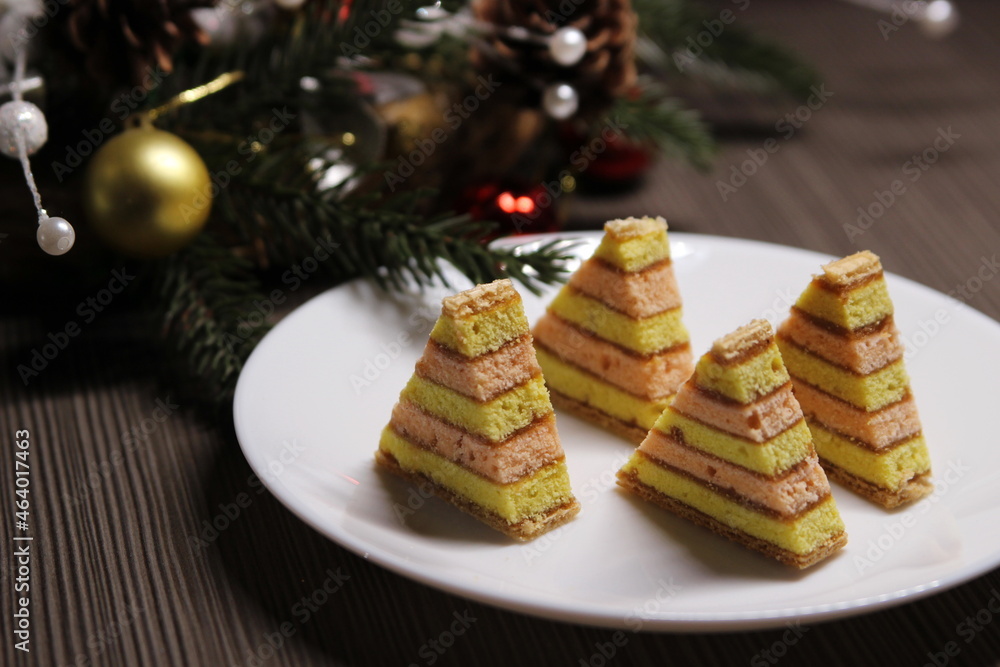 Christmas tree cookies on a white plate, a Christmas tree branch decorated with red and yellow balls on a dark wooden background. New Year's still life with cookies. Christmas tree cookies on a white plate, a Christmas tree branch decorated with red and yellow balls on a dark wooden background. New Year's still life with cookies.