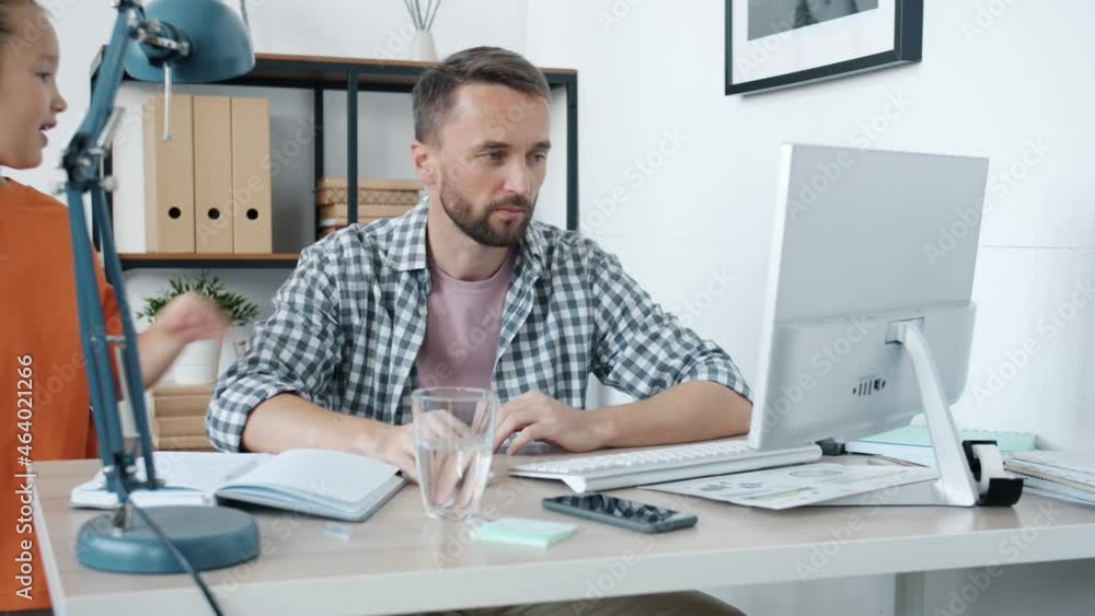 Hyperactive child is bothering annoyed father who is working from home using computer and talking on mobile phone sitting at desk in light room Hyperactive child is bothering annoyed father who is working from home using computer and talking on mobile phone sitting at desk in light room