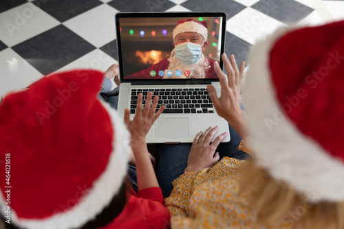 Caucasian mother and child in santa hat on christmas laptop video call with santa claus in face mask