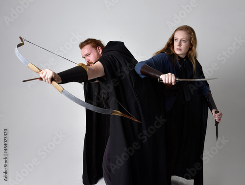 Full length portrait of red haired couple, man and woman wearing medieval viking inspired fantasy costumes, standing fighting pose holding archery bow and arrow and long sword weapons, isolated on 