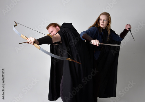 Full length portrait of red haired couple, man and woman wearing medieval viking inspired fantasy costumes, standing fighting pose holding archery bow and arrow and long sword weapons, isolated on 